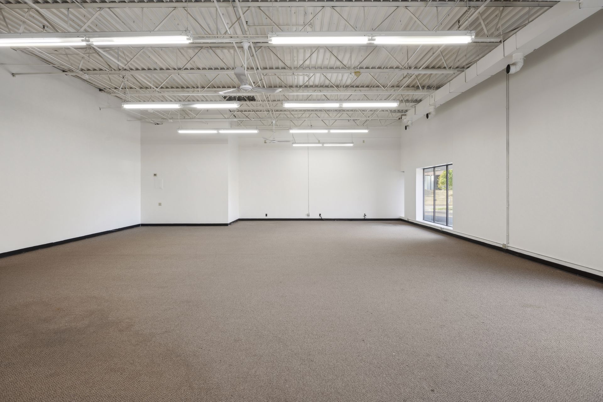 Large open carpeted suite with white walls, exposed painted ceiling structure and a storefront window