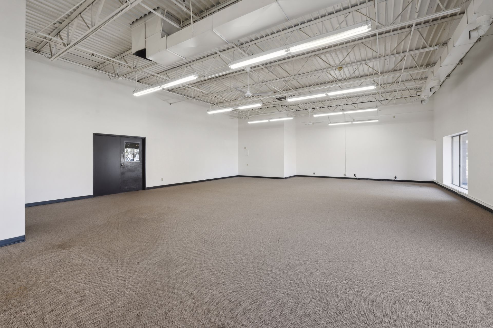 Large open carpeted suite with exposed painted ceiling structure and a door in the side wall