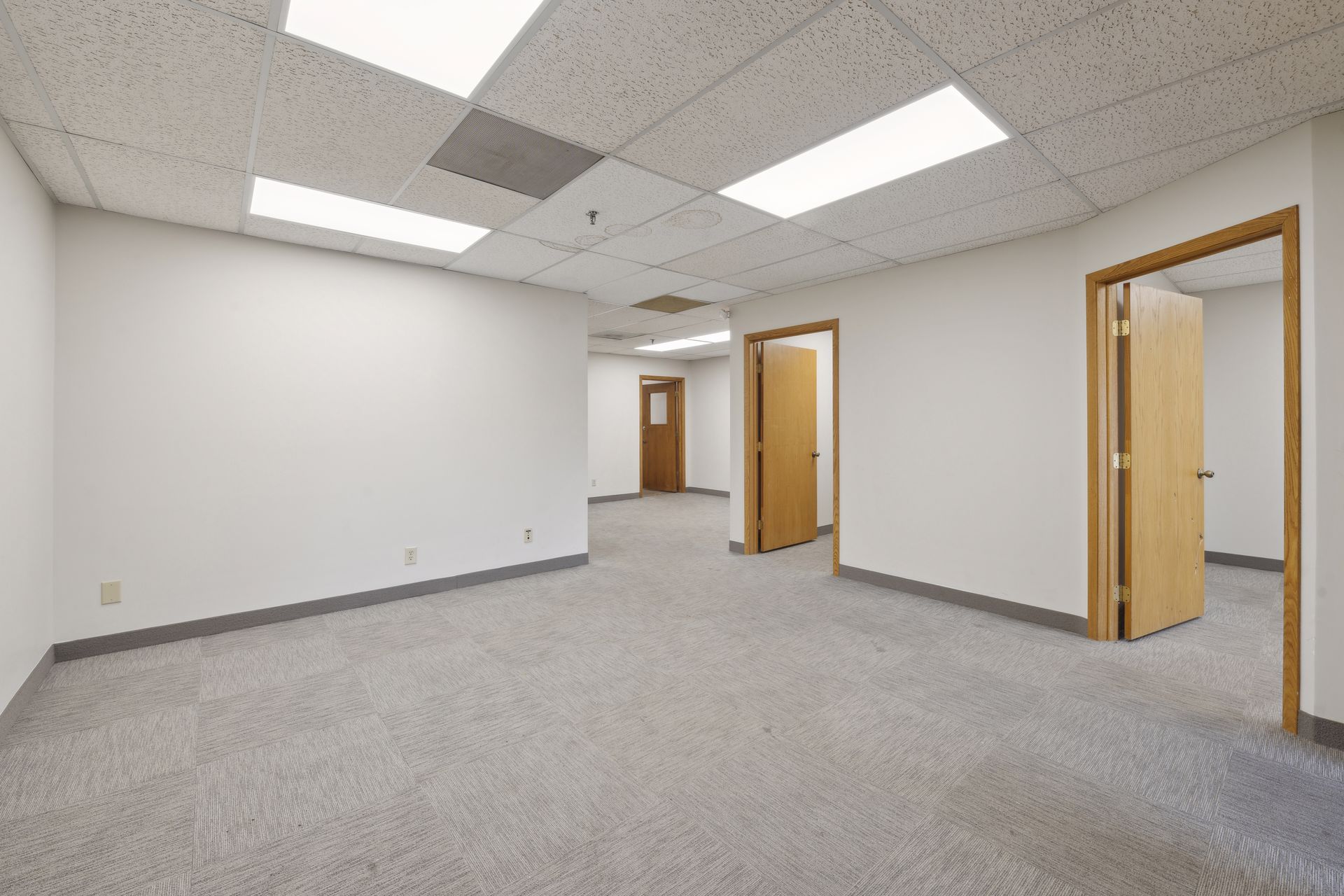 Empty carpeted office room with a suspended tile ceiling and two wood doors opening to further rooms