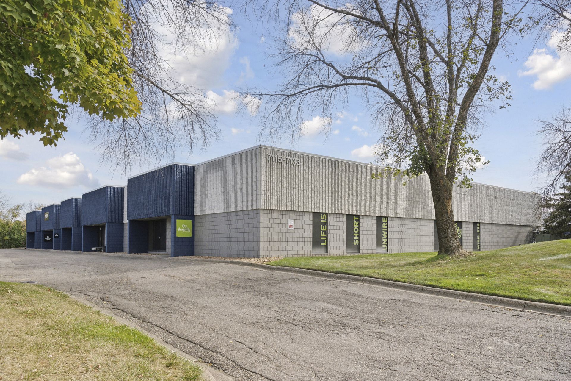Angled view of a single-storey blue and grey commercial building framed by a mature tree