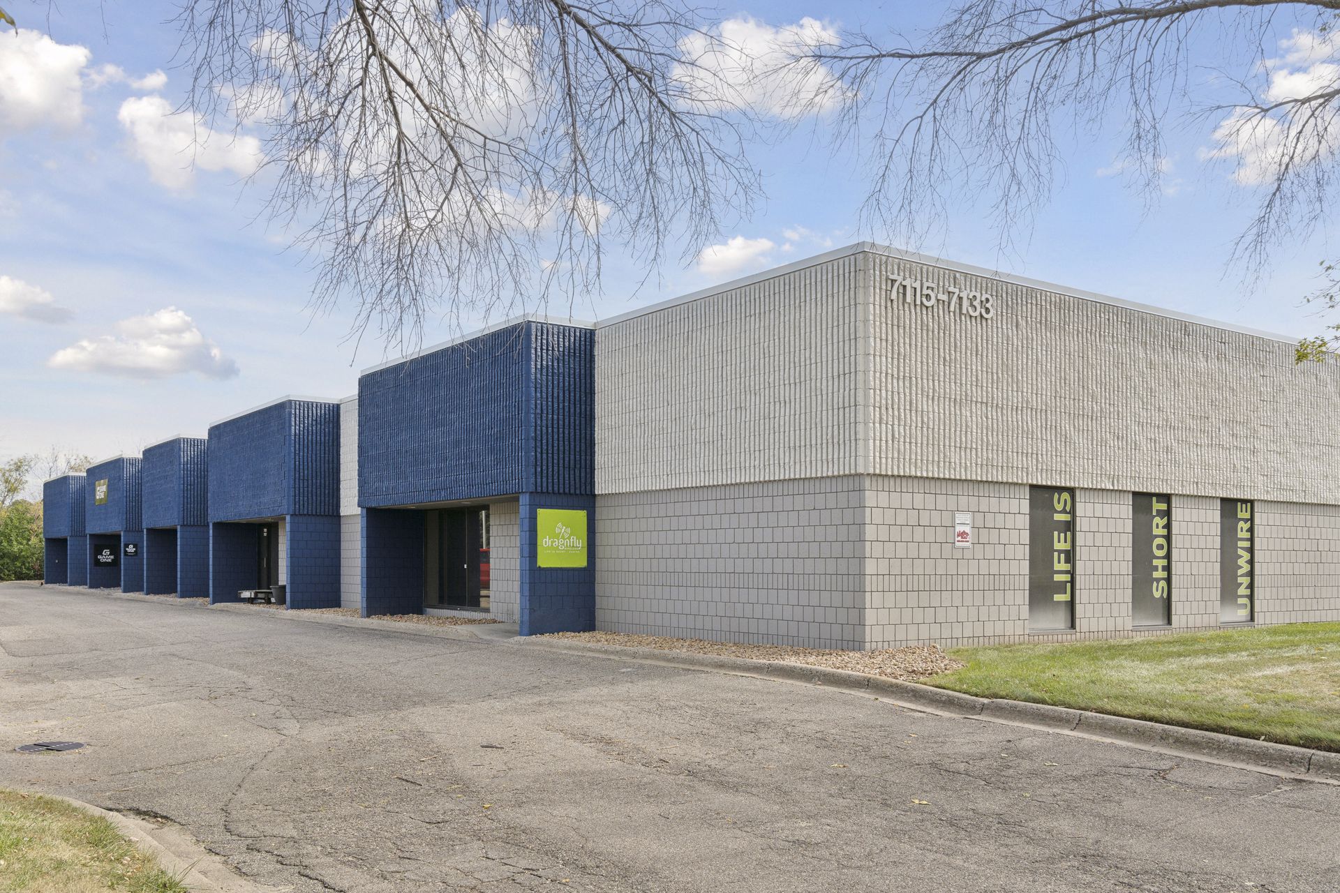 Corner of a single-storey blue and grey block building with the address numbers 7115-7133 on the end wall