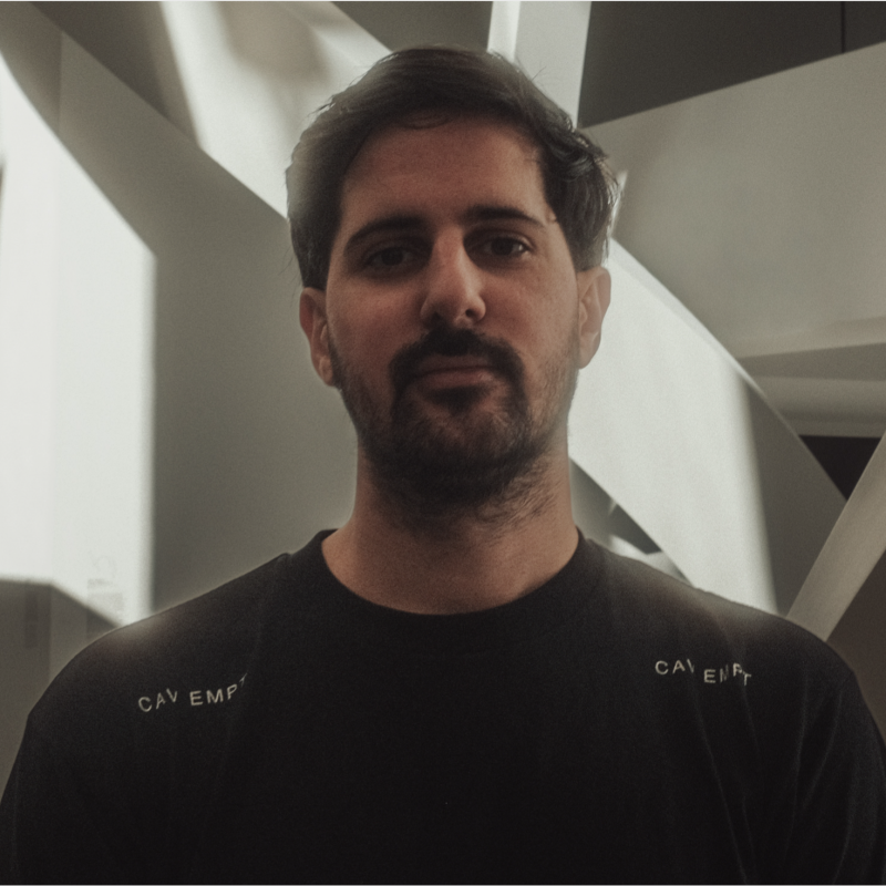 Homme avec cheveux bruns et barbe courte, portant un t-shirt noir avec le texte partiel "CAV EMPT" visible sur les épaules, devant un arrière-plan abstrait aux formes angulaires blanches et ombres.