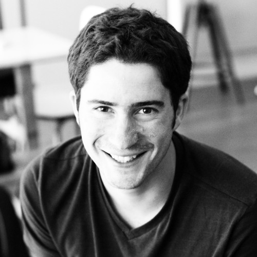 Portrait en noir et blanc d'un homme souriant aux cheveux courts, portant un t-shirt à col en V, avec un fond intérieur flou.