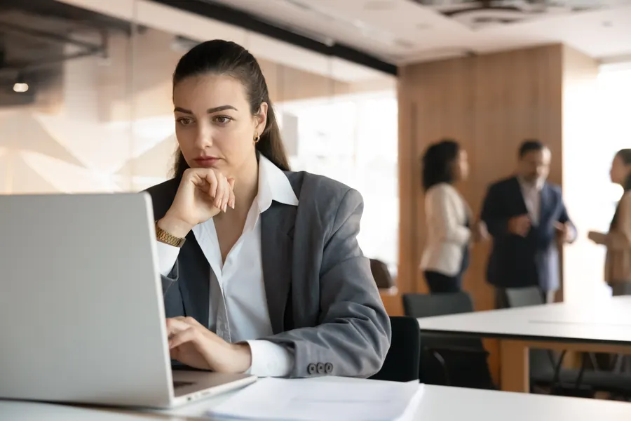 Professional reading a message on a laptop with a concerned expression while colleagues talk in an open office