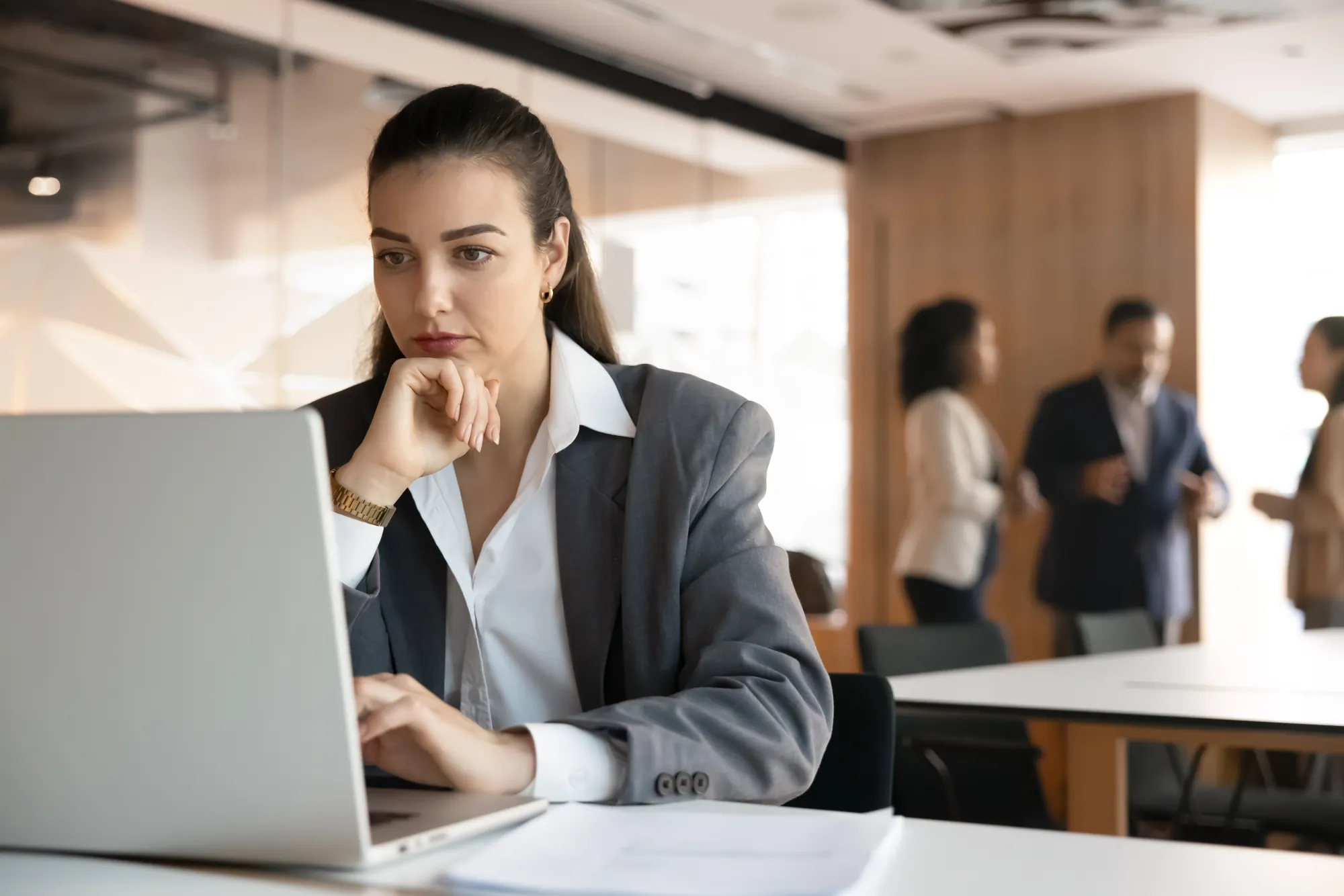 Professional reading a message on a laptop with a concerned expression while colleagues talk in an open office