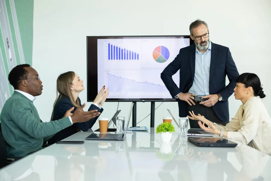 Leader presenting bar and pie charts on a screen while colleagues around a conference table discuss the results