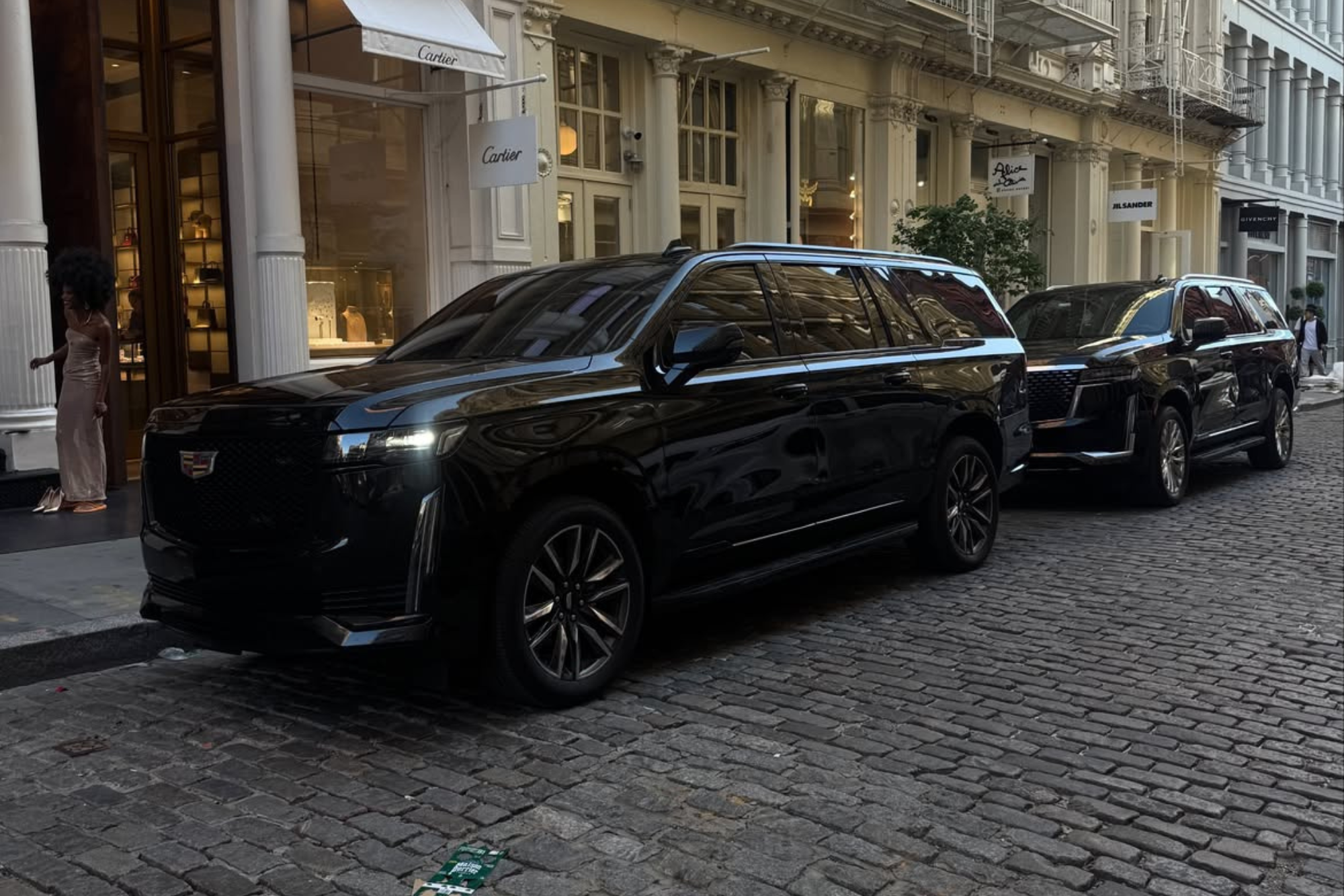 Two black Cadillac SUVs parked on cobblestone street in front of luxury stores and a woman in a dress.