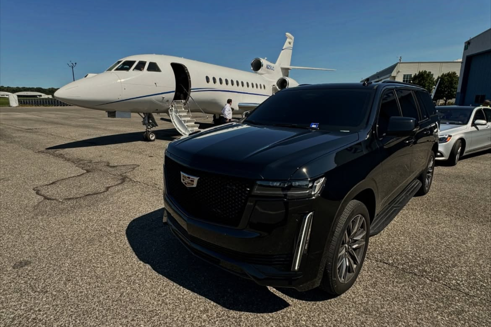 Black Cadillac SUV parked on tarmac near private jet with open door and stairs.