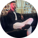 Man in black jacket and white pants sitting relaxed on a black couch in a stylish room.