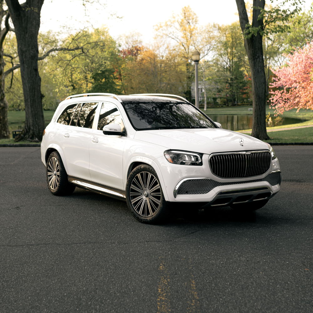 White luxury Mercedes-Benz SUV parked on street with trees and pond in background.
