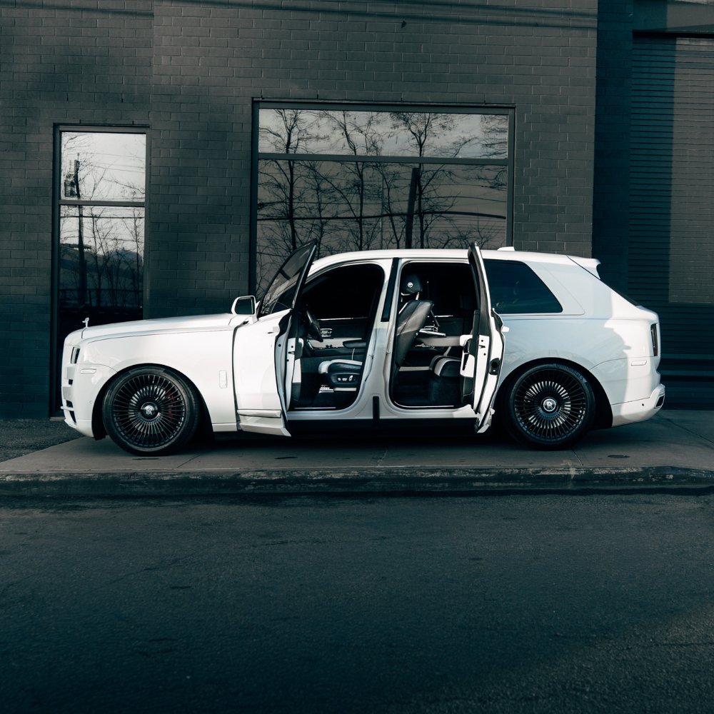 White luxury SUV with front and rear doors open parked by a dark brick wall with tree reflections in windows.