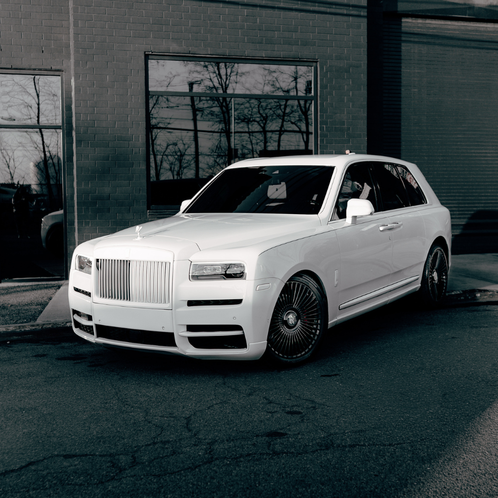White Rolls-Royce luxury SUV parked on a cracked asphalt road near a brick building.
