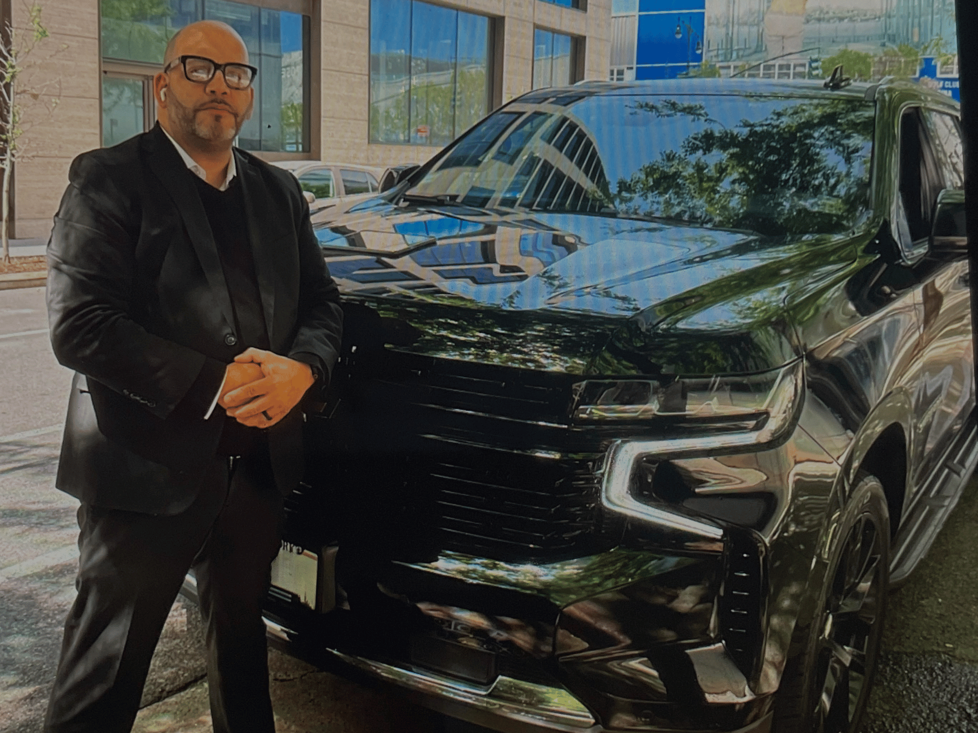 Man in black suit and glasses standing beside a shiny black SUV parked on a city street.