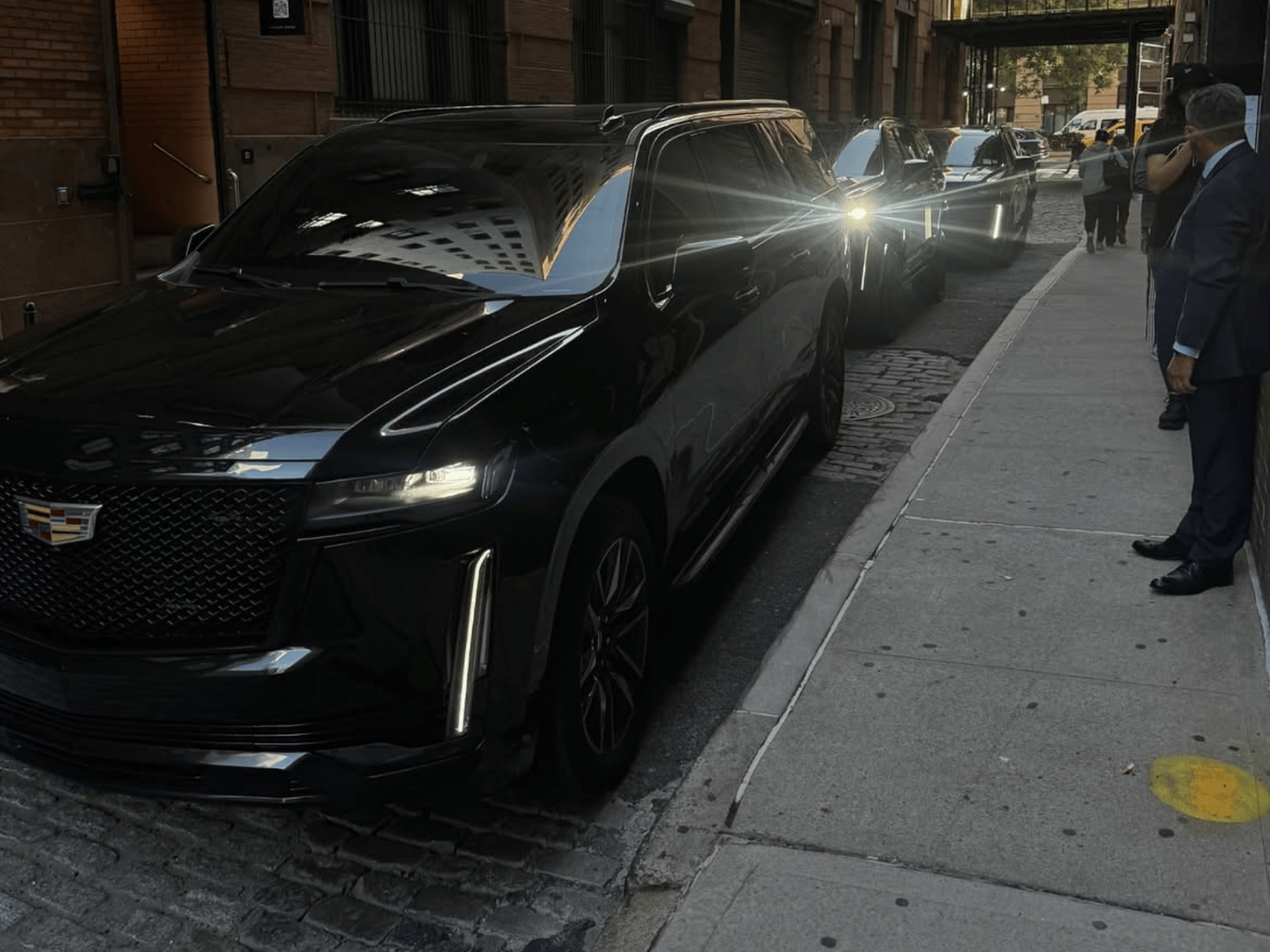 Three black Cadillac SUVs parked on a city street with people standing on the sidewalk.