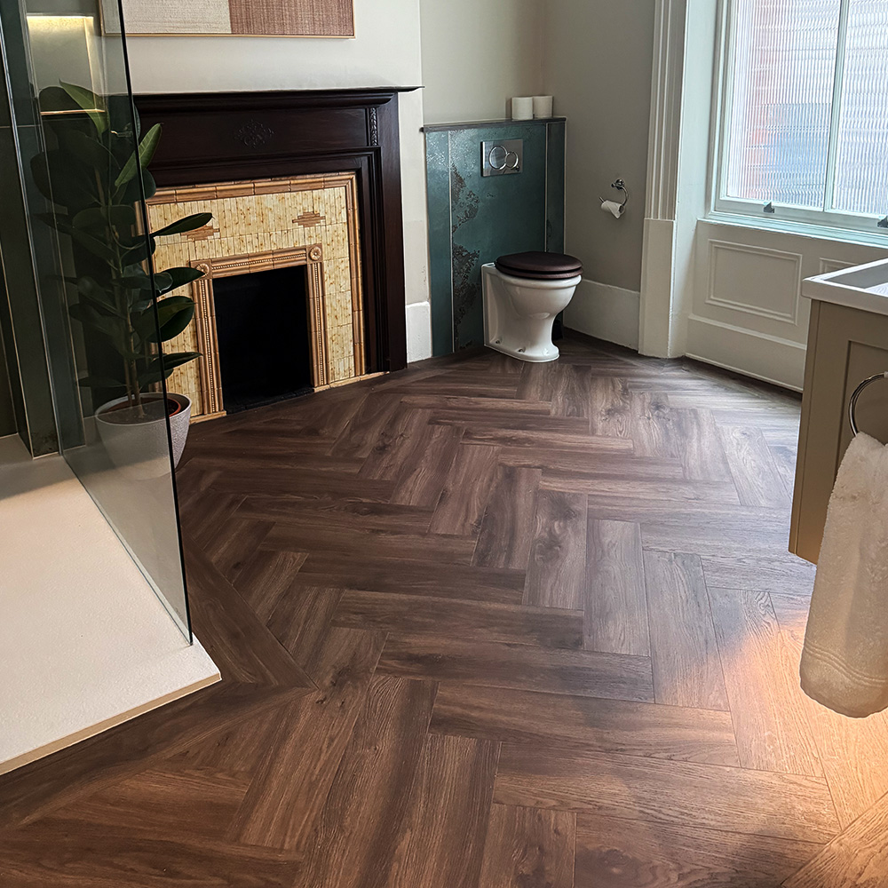 Bathroom with herringbone wood floor, white toilet with dark seat, vintage decorative fireplace, large frosted window, towel ring with white towel, and shower partially visible with glass wall and potted plant inside.