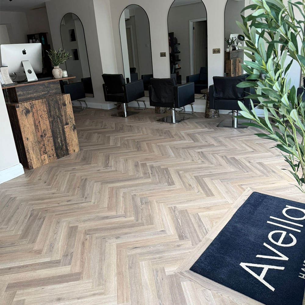 Interior of a modern hair salon with light herringbone wood flooring, three black styling chairs in front of tall arched mirrors, a rustic wooden reception desk with a computer, and a dark blue mat with white lettering partially visible, alongside a leafy green plant on the right.