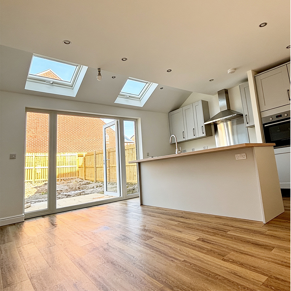 Modern kitchen with wooden floors, white cabinets, stainless steel oven and hood, large island with sink, and glass doors opening to a fenced backyard with brick house visible.