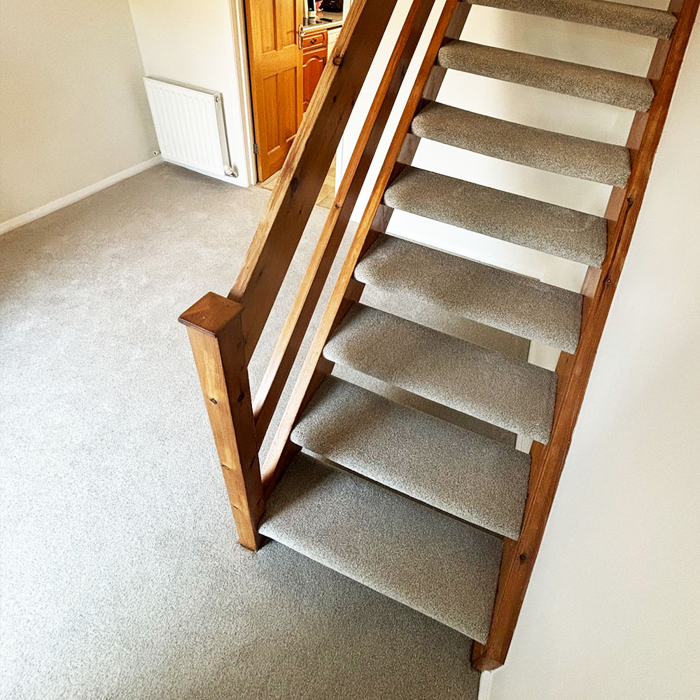 Carpeted staircase with wooden handrails inside a beige-walled room with gray carpet floor and an open wooden door leading to another room.