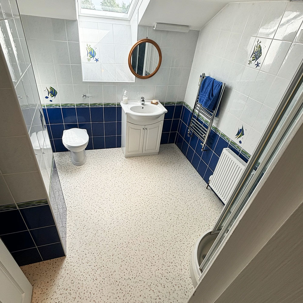 Small bathroom with white and blue tiled walls decorated with fish tiles, a white sink cabinet, white toilet, round wooden-framed mirror, wall-mounted towel rack with a blue towel, and a corner shower enclosure with sliding doors.