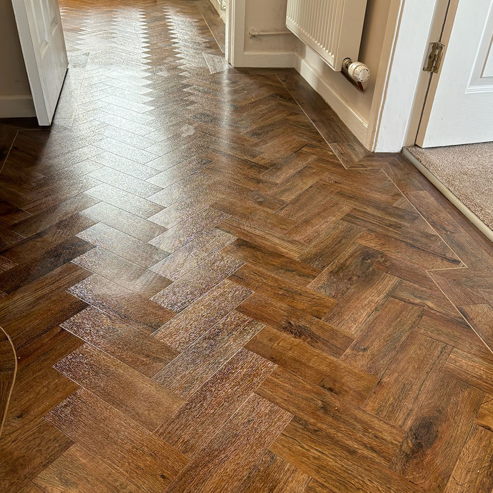 Indoor floor with brown herringbone patterned wood laminate or vinyl flooring extending through a hallway with two white doors partially visible on the sides and a wall-mounted radiator on the right.