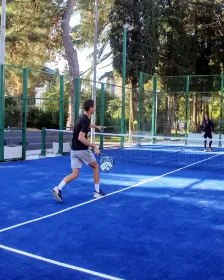 Tennis players practicing on an outdoor blue court surrounded by green fencing and trees.
