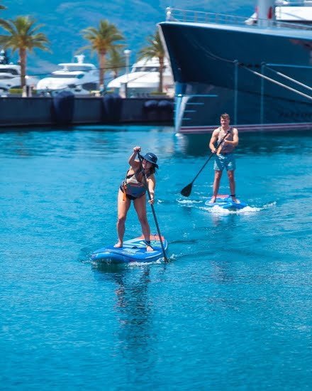 Yacht docked at marina with paddleboarders enjoying water activities.