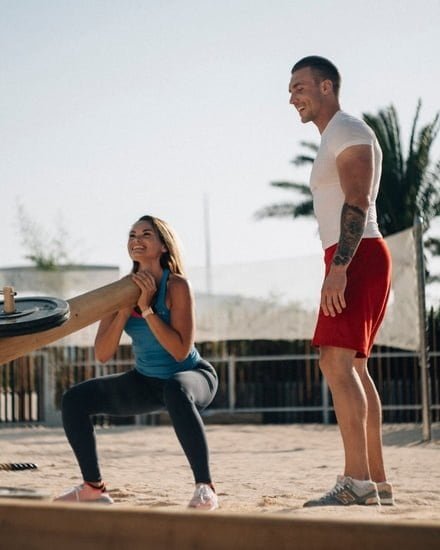 Outdoor strength training with a woman performing a squat with a heavy log, supervised by a man.