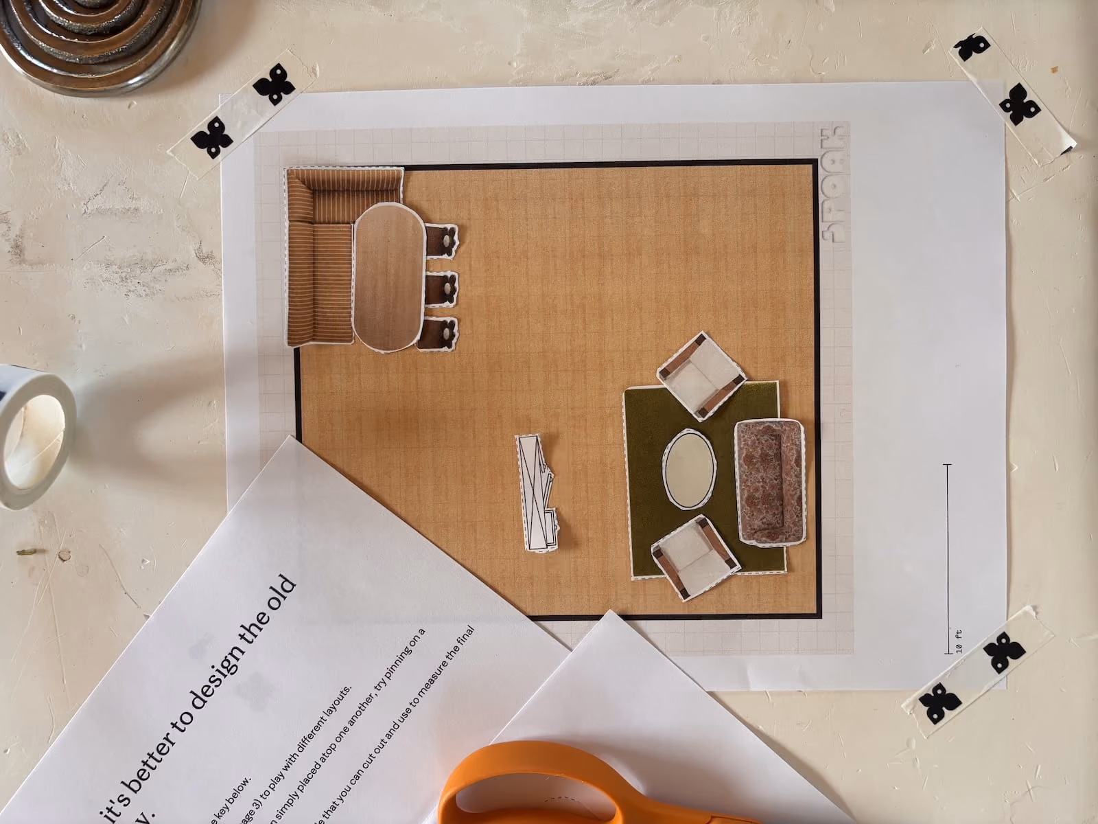 Top-down view of printed floor plan taped to wall with paper cutouts of furniture and orange scissors nearby.