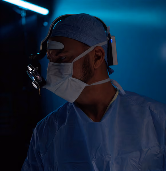 Surgeon wearing blue scrubs, face mask, surgical headlamp, and cap in a dimly lit operating room.