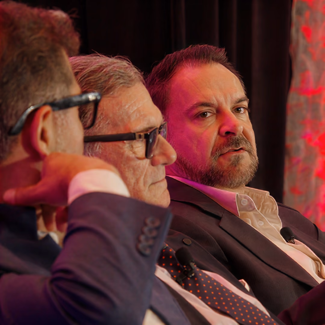 Three men in suits with microphones engaged in a panel discussion under red lighting.
