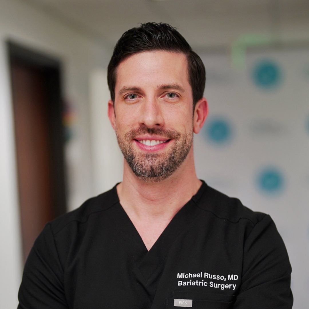 Smiling man in black medical scrubs with Michael Russo Bariatric Surgery embroidered on chest.