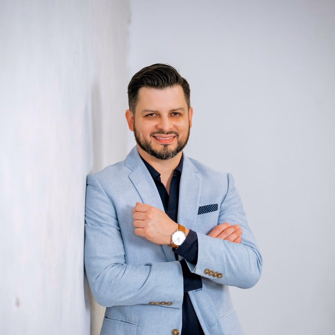 Man with dark hair and beard wearing a light blue blazer and watch, smiling with arms crossed.