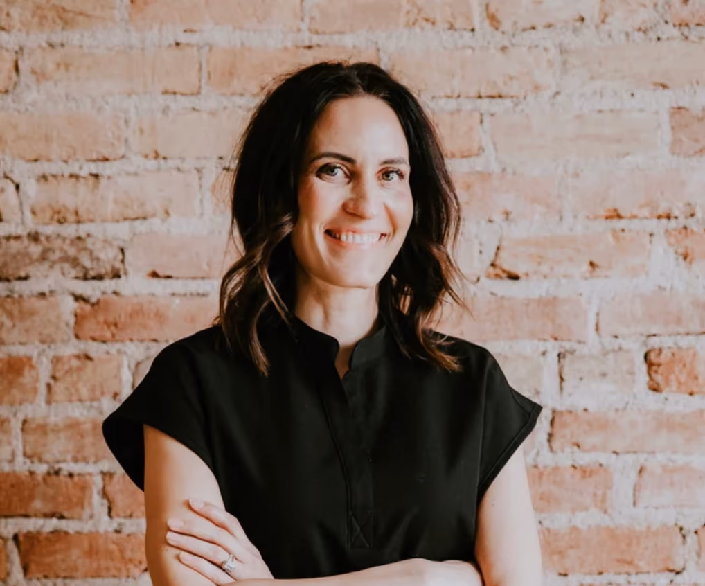 Smiling woman with dark hair and a black shirt standing with arms crossed against a brick wall.
