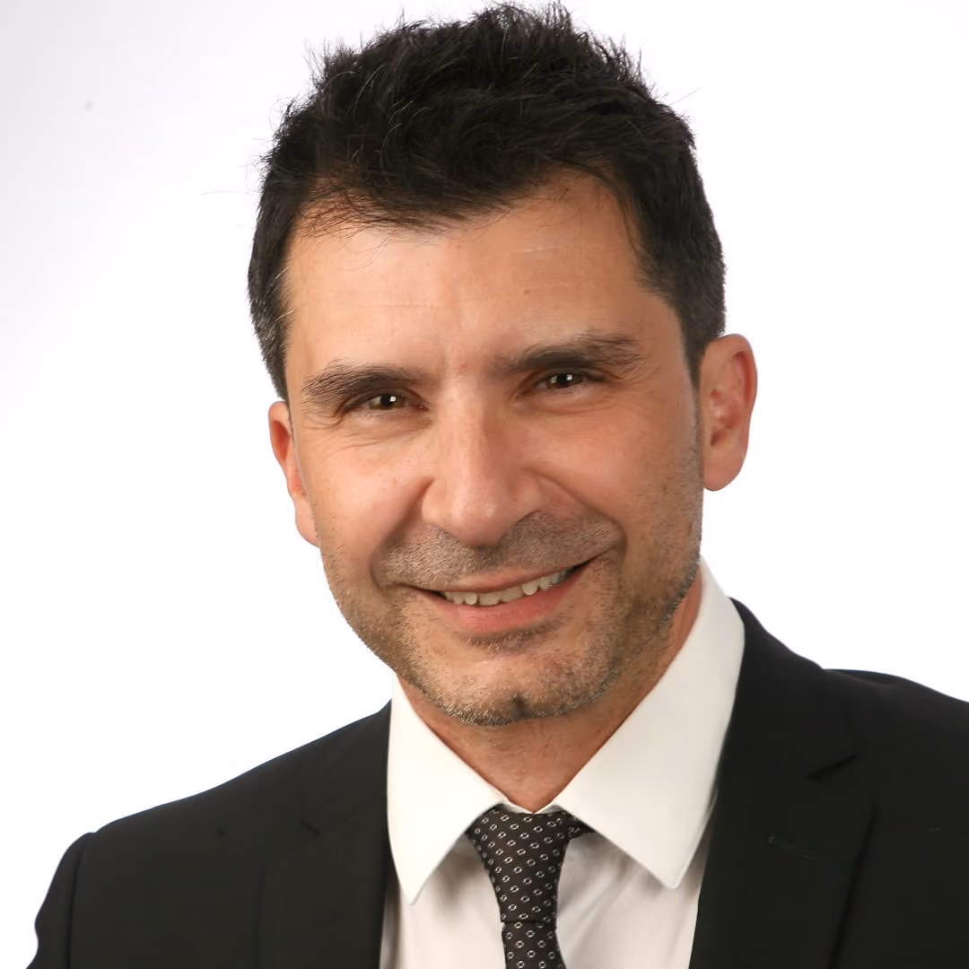 Smiling man with short dark hair in black suit, white shirt, and patterned tie against white background.