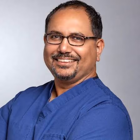 Smiling man with glasses wearing a blue scrub top against a gray background.
