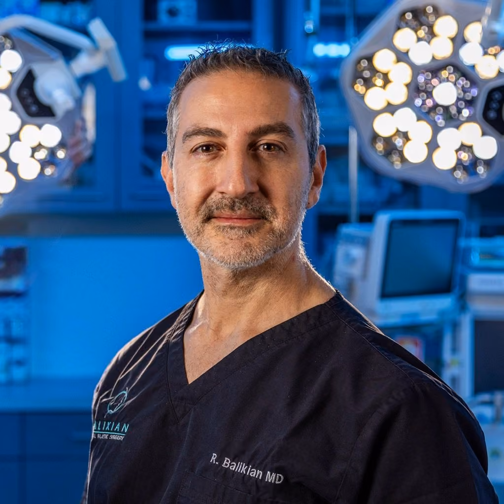 Middle-aged male doctor in black scrubs in a surgical room with equipment and bright lights behind him.