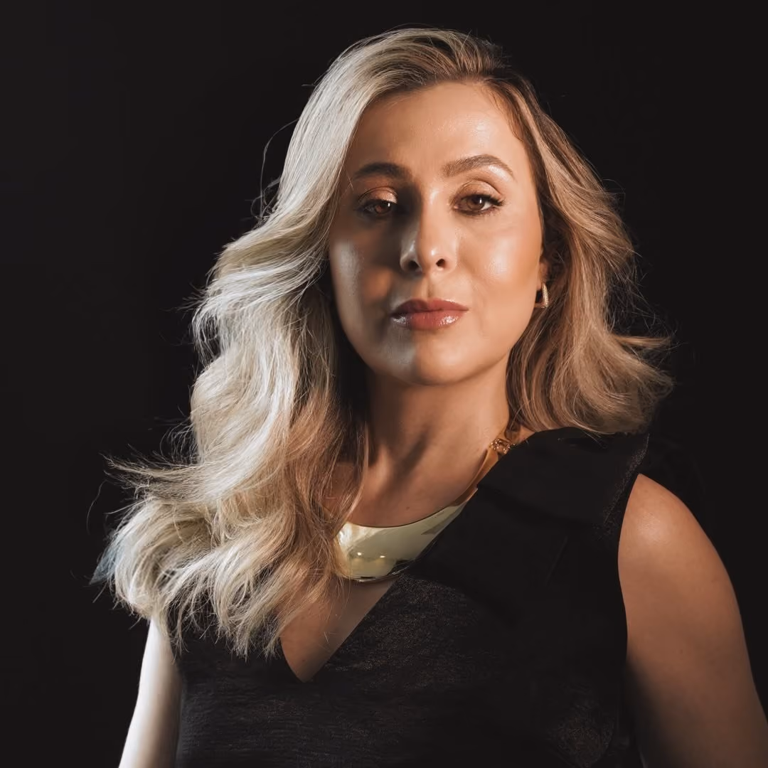 Confident woman with wavy blonde hair wearing a black top and gold necklace on black background.