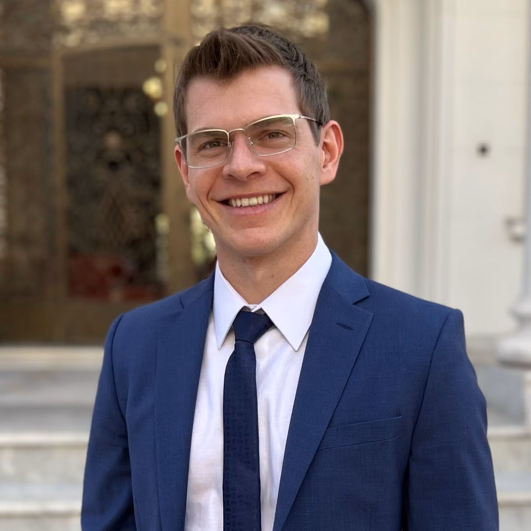Smiling man wearing glasses, a blue suit, white shirt, and dark tie standing outdoors in front of steps.
