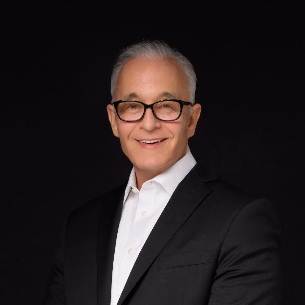 Smiling older man with gray hair, glasses, white shirt, and black suit jacket on black background.