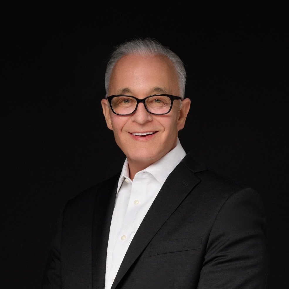 Smiling older man with gray hair, glasses, white shirt, and black suit jacket on black background.