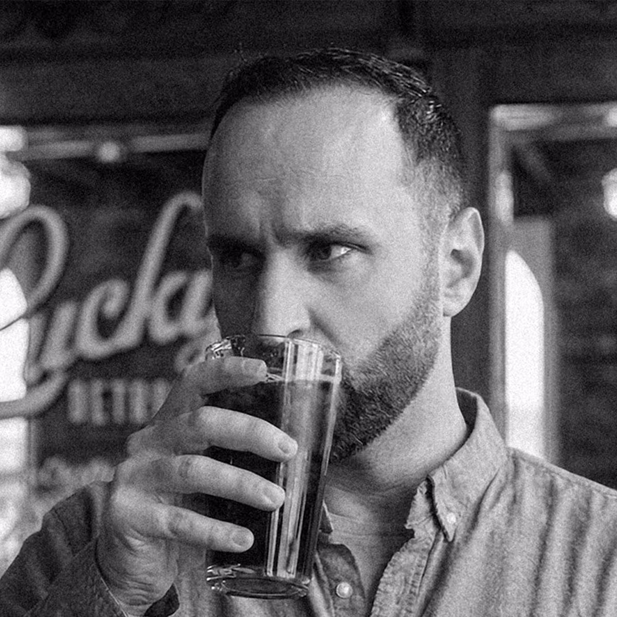 Black-and-white photo of a man with a short beard and cropped hair, wearing a button-up shirt, sipping from a glass at a coffee shop, with vintage-style signage reading "Lucky Detroit" blurred in the background