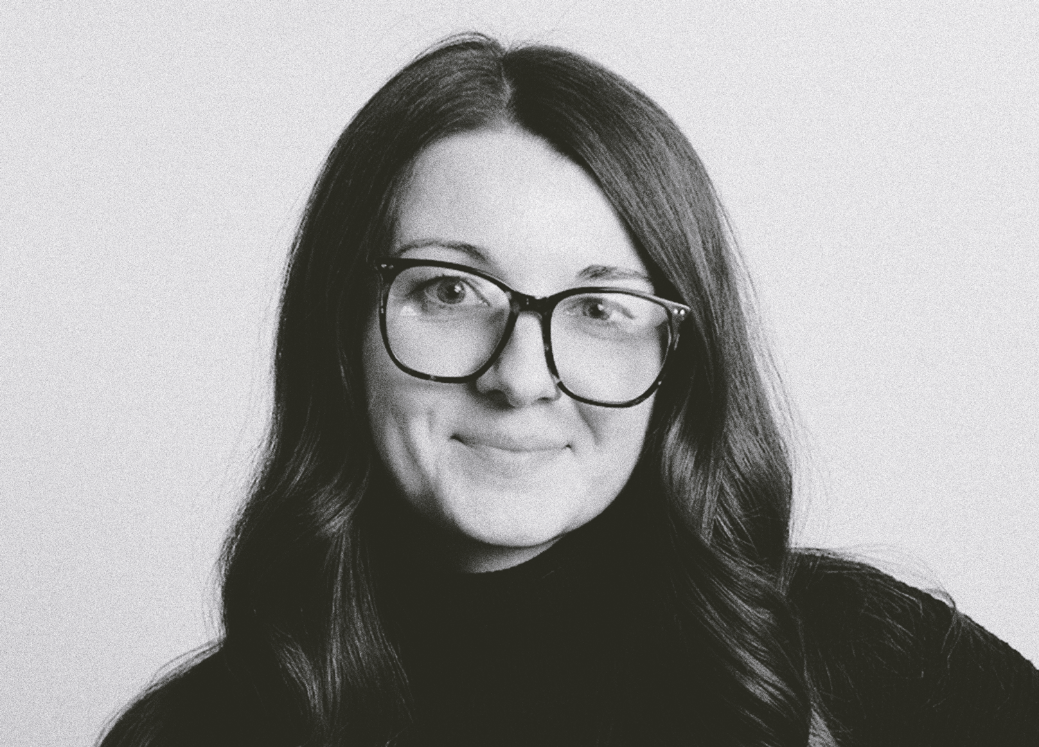 Portrait of a woman with long wavy hair wearing glasses and a dark top, smiling against a clean neutral background