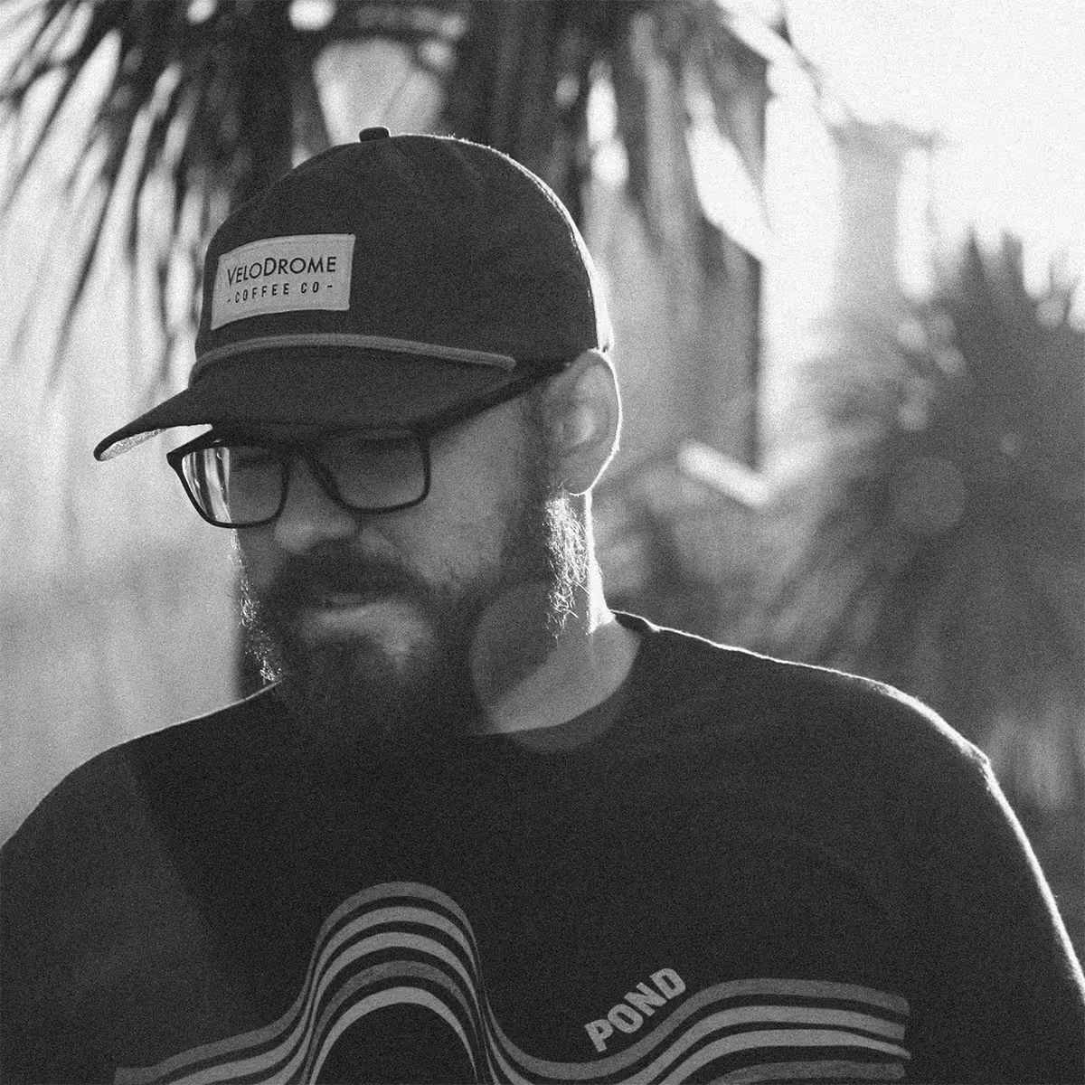 Black and white portrait of a man with glasses, beard, and a cap, looking downward outdoors
