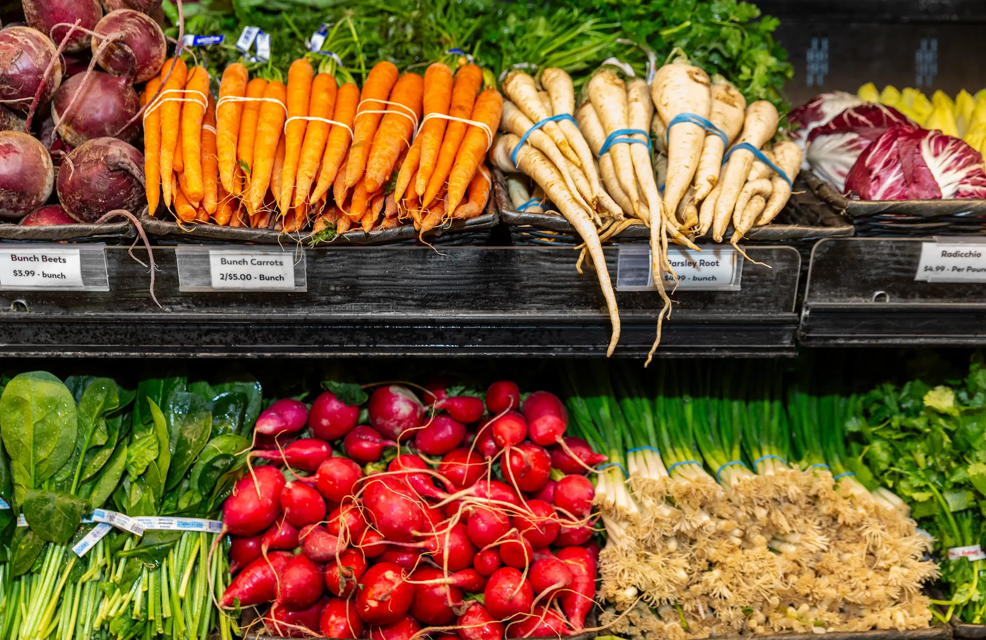 Fresh produce display at Western Market with labeled price signs for bunch beets, bunch carrots, and parsley root on the top shelf, plus radicchio, spinach, radishes, and green onions below.