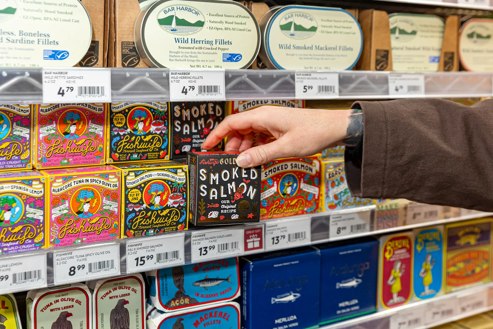 A hand reaching for a box of Fishwife Gold Smoked Salmon on a shelf stocked with tinned seafood, including Bar Harbor sardines and mackerel, and colorful Fishwife tuna and salmon boxes with price tags.