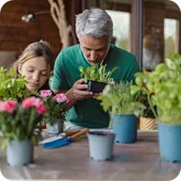 An older man and a young girl examining potted plants on a wooden table indoors.