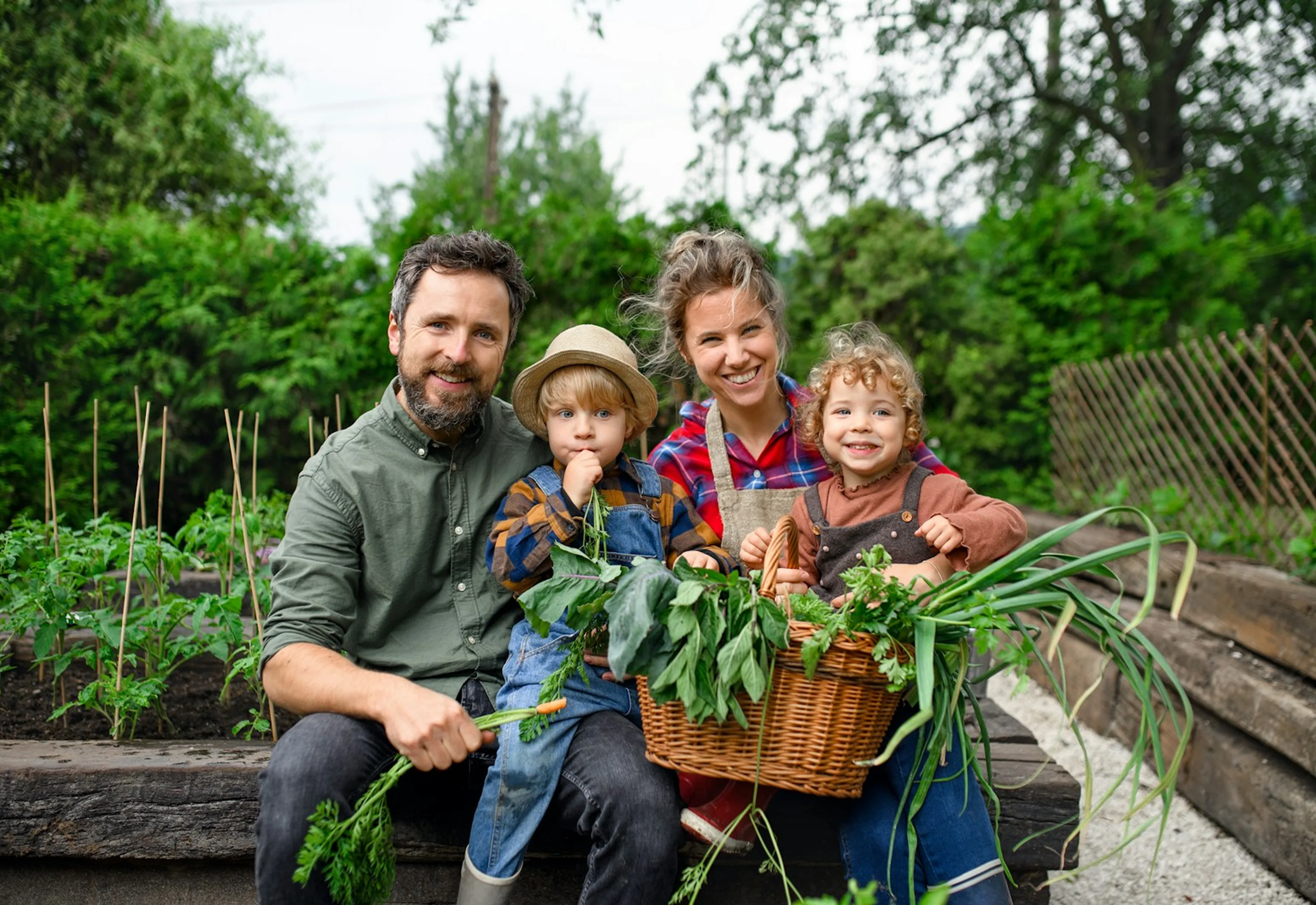 Happy family of four sitting in a garden holding a basket and fresh vegetables, smiling at the camera.