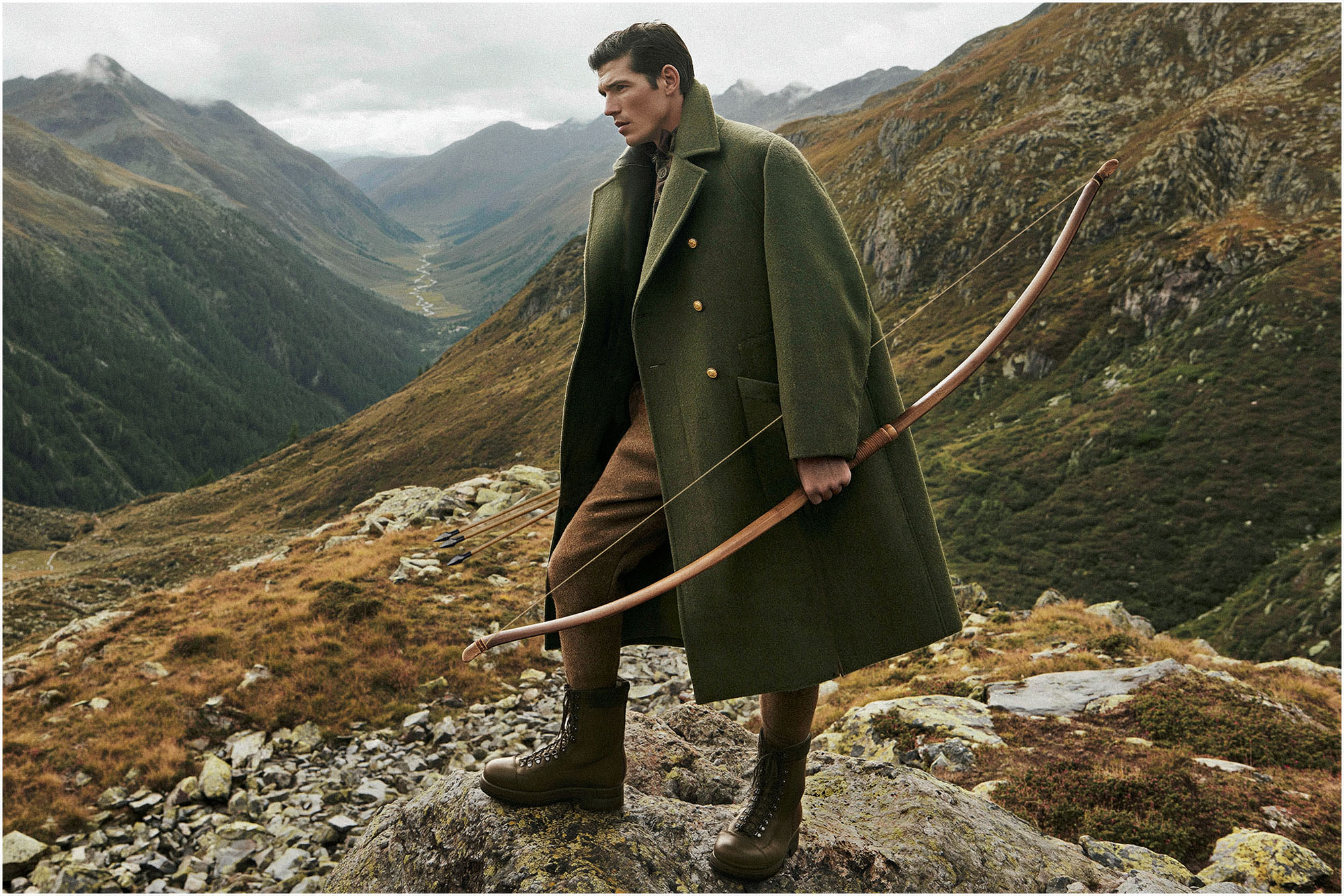 Fashion editorial portrait of a man wearing a dark green overcoat and brown trousers, holding a wooden bow in a rocky alpine mountain landscape in Val Tantermozza, Switzerland.