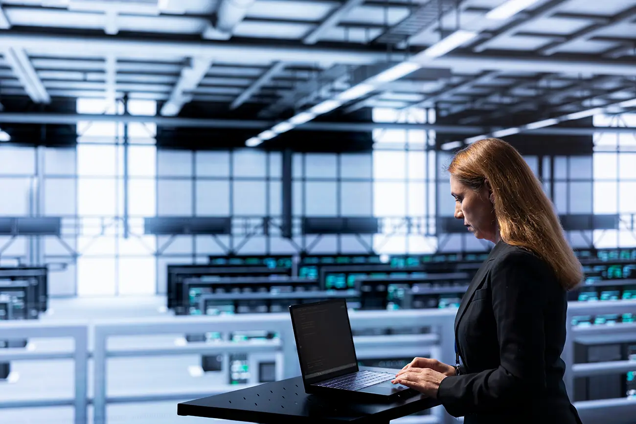 Woman working on a laptop in a large, modern office space with multiple desks and screens.