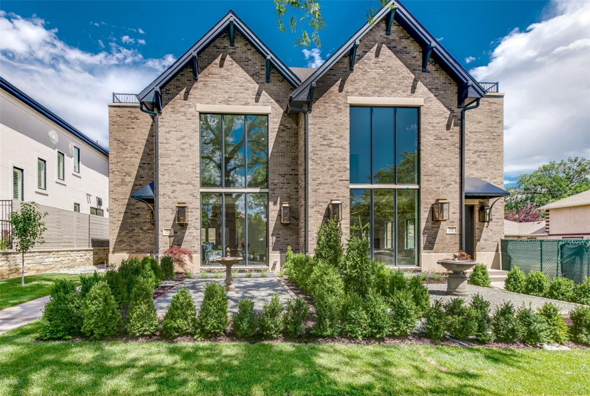 Modern brick duplex with large floor-to-ceiling windows, slate roof accents, and a neatly landscaped front yard featuring trimmed shrubs, two stone water fountains, and a green lawn under a blue sky with scattered clouds.